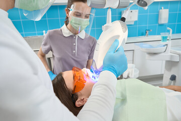 Dentist carrying out teeth-whitening procedure supervised by female nurse