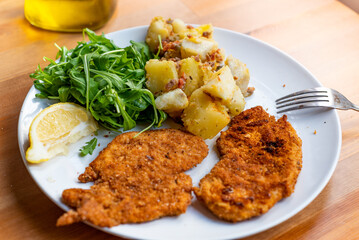 milanesa steak or breaded schnitzel served on dish with green salad and cooked potato salad on wooden table. homemade simple lunch