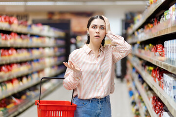 High price and inflation. Portrait of young shocked caucasian woman holds cart and takes hand to...