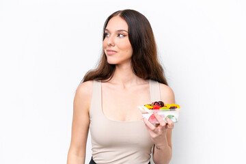Young caucasian woman holding a bowl of fruit isolated on white background looking to the side and smiling