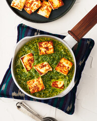 Bright green palek panner in a pot on patterned trivet with pan fried paneer.