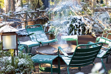 Winter garden in the snow with chairs around a fire. Smoke from the campfire.