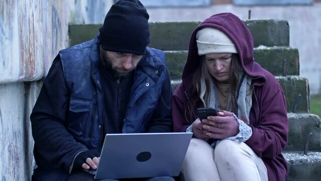 Two frozen homeless people, a man and a woman, sit in the street. They are holding a laptop and a mobile phone. The couple are looking for job advertisements.