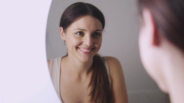 Happy positive young woman looking at her reflection at the mirror standing in bathroom and smiling, satisfied with her appearances, spent beautiful night, feeling sensual and sexual