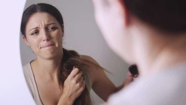 Sad young woman looking at her reflection at the mirror combing her long brown hair, dissatisfied with splited and dry ends, need to change care shampoo and hair conditioner