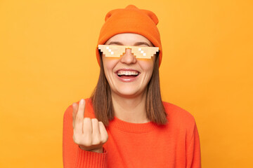 Portrait of delighted happy optimistic woman wearing orange jumper, beanie hat and glasses in minecraft style, showing korean love mini gesture, standing isolated over yellow background.