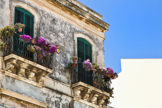 Facade of a residential building with shutters and balconies with blossoming plants; Syracuse, Sicily, Ortigia, Italy