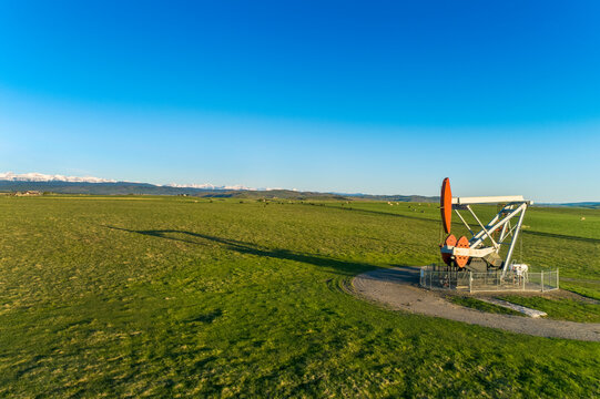 Pumpjack in a green field with long shadow under a blue sky and snow-covered mountain range in the background, West of Longview; Alberta, Canada