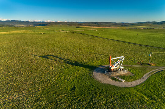 Aerial view of a pumpjack in a green field with long shadow under a blue sky and snow-covered mountain range in the background, West of Longview; Alberta, Canada