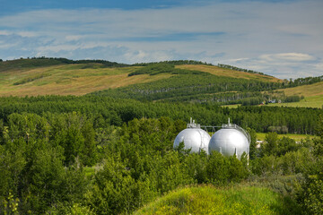 Two spherical white oil storage tanks amongst trees and hills with clouds and blue sky; Turner Valley, Alberta, Canada