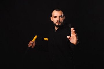 Young serious bearded man practices nunchaku martial art in a dark black studio.