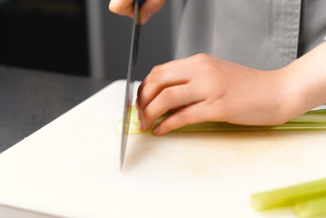 Hands of a female chef are cutting fresh green celery on a plastic board. Side view.