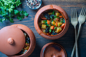 Stewed potato, carrot, onion, tomato and prunes in a clay pots with lid on wooden background, closeup