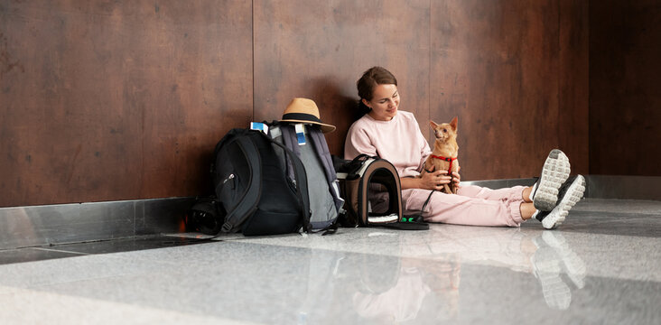 Young adult woman travelling with her small dog sitting on floor at airport terminal while waiting for boarding.