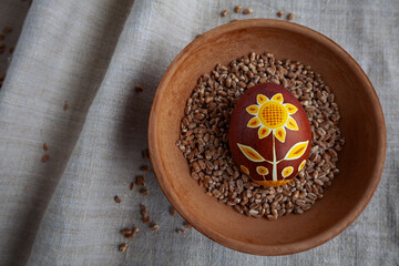 Ukrainian Easter egg in brown-yellow colors with a sunflower flower on a background of linen fabric in a plate with grain