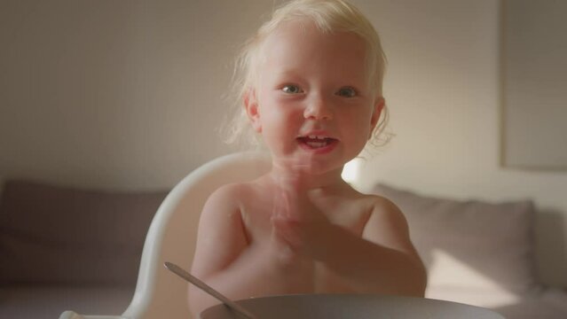 Blonde funny cute girl boy little pretty infant child sit at small kitchen table applauding clap hands happy newborn kid daughter or son eat porridge breakfast tasting food childhood innocence concept