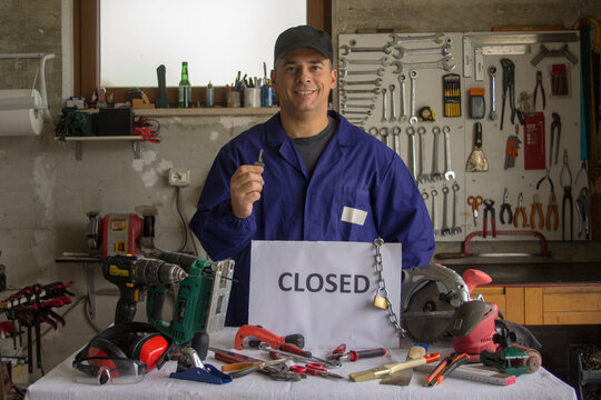 Image of a smiling craftsman in his workshop holding a wrench in his hand and in front of a bench with tools and a closed sign. Reopening of business.
