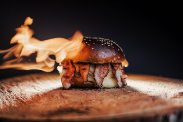 
Tasty beef, cheese, burger with many slices of bacon, warm coming out of smoke on a wooden cutting board on a black background, 
with fire on the stage.  - obrazy, fototapety, plakaty