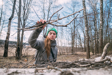 Preparation for the heating season, firewood harvesting in rural areas. A young woman in warm clothes collects sawn wood and branches in the forest on a sunny day. © Anna