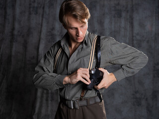 Fototapeta premium A lawyer with a pistol in a holster, gray shirt and trousers with suspenders, retro style. A young man poses in the studio on a gray background