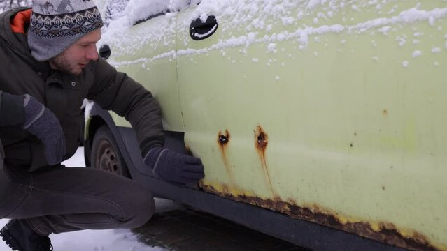 A man shows rust on a car door from winter reagents. Close-up, selective focus on rust and hand.