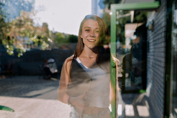 Happy smiling European girl with happy emotions is posing at camera through the window near modern cafe in the city . Girl is holding laptop and preparing for work remote