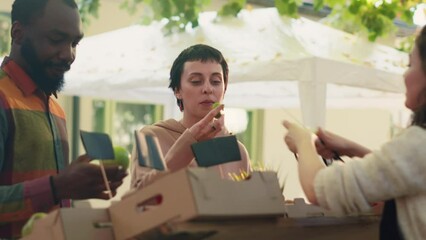 Female farmer giving free slices of apples for clients to taste, diverse people enjoying product samplingg at food market. Young woman giving samples of organic produce to cusomers. Handheld shot.