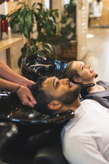 Indian male client having his hair washed by hairdresser next to blonde female customer in a professional hair salon. High quality photo 