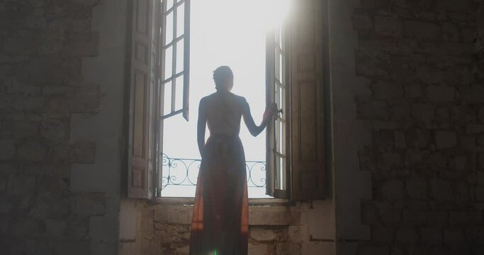 Princess in beautiful dress looks through open window in her castle, back view.