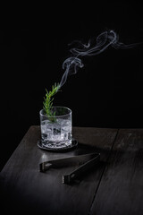 Rosemary branch lit with smoke inside a mezcal drink with tequila on ice, ice tongs on a wooden table