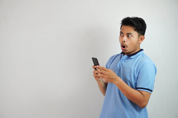 shocked young asian man holding phone and open mouth wearing blue t shirt isolated on white...