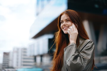 Portrait of a woman business talking on the phone with a smile with teeth and red hair in front of tall buildings in the city, the concept of technology and business online