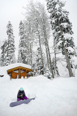 A snowboarder in a mountain forest near a hut covered with snow in winter in a snowstorm