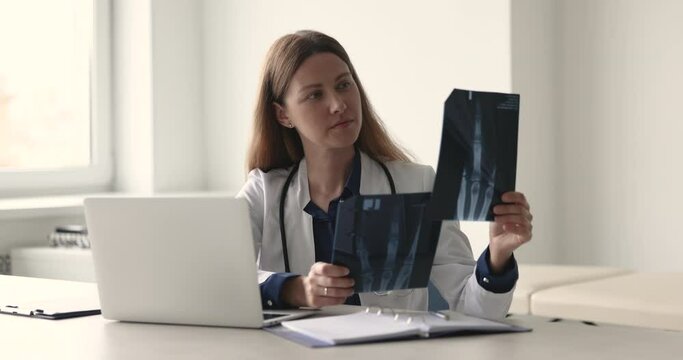 Focused woman radiologist in white coat, female doctor specializing in radiology sit at desk holding digital images reviewing, analyze x-rays results. Professional medical worker workflow at workplace