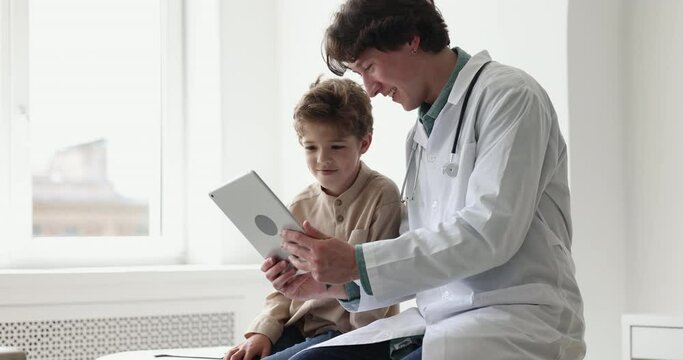 Young male pediatrician talks to little boy, explain treatment plan or builds trusting friendly relationships showing to small patient new game or amusing app on tablet. Medical services, modern tech