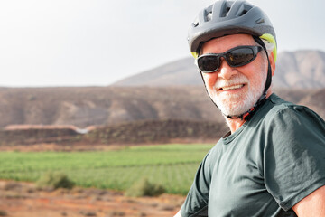 Happy cyclist senior man with protective helmet riding bicycle in the countryside on a sunny day. Handsome bearded male enjoying healthy lifestyle and sport activity