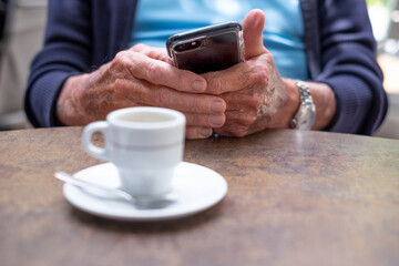 Fototapeta na wymiar Closeup on old senior man hands using mobile phone. Elderly caucasian male sitting at cafe table with an espresso coffee cup while looking at his smartphone