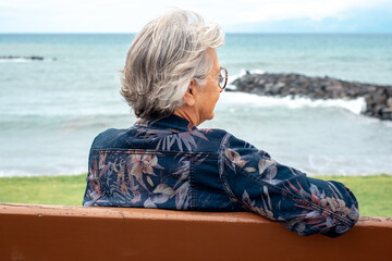 Back view of senior woman sitting on a bench at the beach in a winter day enjoying free time vacation or retirement. Trendy gray haired lady with eyeglasses relaxed in front to the sea