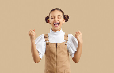 Happy child is happy to succeed and win. Joyfully excited prteen girl happily laughing fists with joy on beige background. Cute caucasian kid girl with close eyes rejoices at goal and shouts cheers.