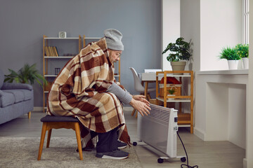 Elderly man wrapped in plaid feeling cold at home. Retired man warming hands on oil radiator having...