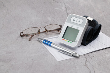 Automatic digital wrist blood pressure monitor, pressure monitoring chart, pen, glasses on gray background
