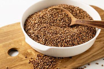 Dry uncooked buckwheat groats in a white dish, wooden board, spoon, on a white background, close-up