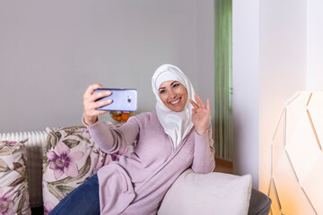 Young Muslim woman having video call via smart mobile phone at home. Happy smiling Muslim woman sitting on sofa, couch and using smart phone at living room at home, learning language, video calling.