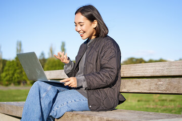 Portrait of young asian woman working remotely from park, sitting on bench with laptop, talking...