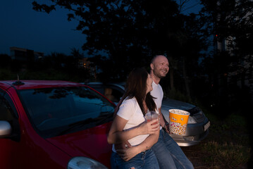 Couple with popcorn standing at the car while watching a movie at drive in cinema. Selective focus. Dating in drive in theater. Entertainment, leisure activities, hobby concept