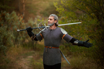 An aged male warrior in chain mail with gray hair and a scar on his face. Knight in armor holds a two-handed sword.