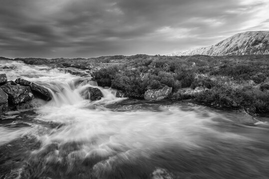 Black and white Majestic Winter landscape image of River Etive in foreground with iconic snowcapped Stob Dearg Buachaille Etive Mor mountain in the background