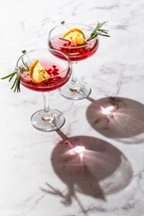 Two wineglass of Pomegranate iced cocktail with rosemary and lemon on white marble background isolated with clear shadows in harsh light. Festive xmas drink, valentines concept