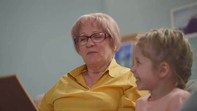 Granny reads fairy tale as actress to happy little child in living room closeup. Grandmother and preschooler girl have fun at home. Family generations