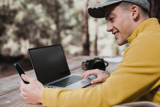 Business or study on fresh air. Happy young protographer man working on laptop and using phone while sitting at the wooden table outdoors in the forest. Concept of smart working in nature